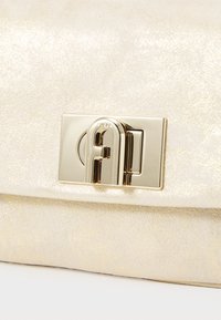 Chiusura tono oro e emblema del marchio su una superficie cremosa e texturizzata. I materiali appaiono lisci con una leggera lucentezza. Design compatto e rettangolare.