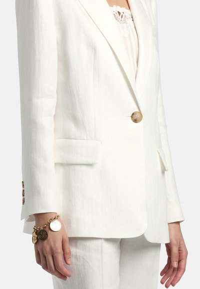 Nő fehér, egygombos, szabott blézerben, hozzáillő fehér nadrágban, csipkés felsőben és arany charm karkötőben a bal csuklóján.