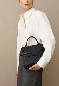 Borsa a mano in pelle nera con un design strutturato, manico superiore e chiusura a patta. Texture liscia e sottili dettagli in hardware. Indossata con una camicia bianca.