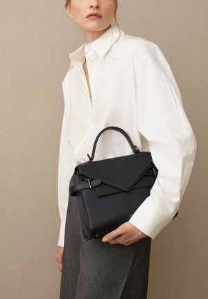 Bolso de mano negro de piel con un diseño estructurado, asa superior y cierre de solapa. Textura suave y sutiles acentos metálicos. Combinado con una camisa blanca.