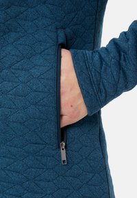 Donkerblauwe gewatteerde stof met een geometrisch patroon, voorzien van een ritszak aan de zijkant en een mouw met een manchet en gestructureerd detail.