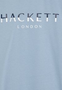Gaiši zils kokvilnas T-krekls ar tekstu "HACKETT" tumši zilā krāsā un "LONDON" baltā krāsā. Gluda tekstūra, apaļa kakla dizains.