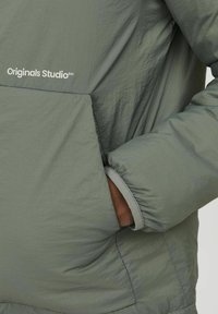 Zielona, lekka kurtka o miękkiej fakturze, z przednią kieszenią i rękawami z mankietami. Na piersi widoczny logo "Originals Studio".