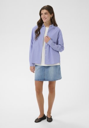 Młoda kobieta o długich włosach, ubrana w luźną, niebieską, paskowaną koszulę na białym topie, dżinsową spódnicę i czarne, siateczkowe baleriny, stojąca na białym tle.