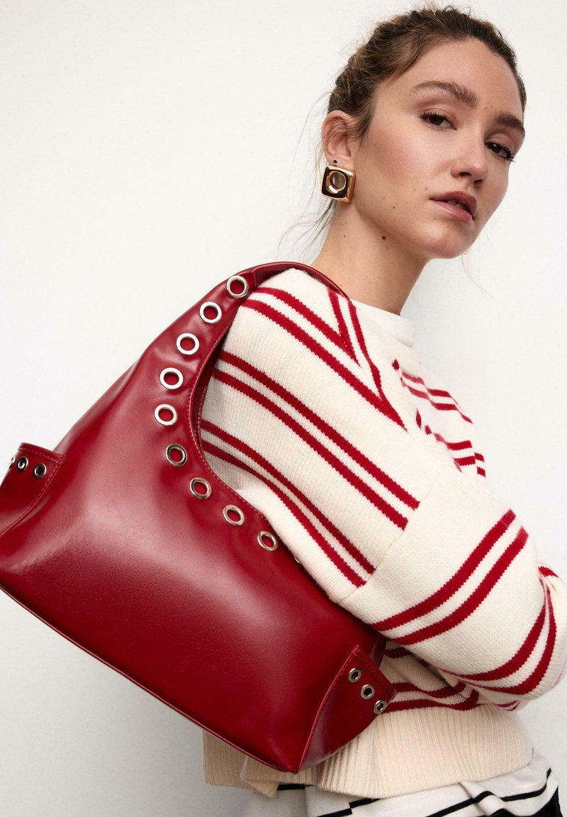 Borsa a mano in pelle rossa con grandi occhielli lungo un lato. Texture morbida e design curvo. La modella indossa un maglione a righe crema e rosso.