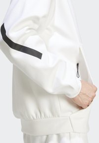 Veste blanche à zip avec une surface texturée, une bande noire sur la manche et des poignets élastiques. Comprend une poche latérale zippée.