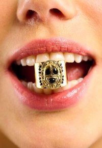 Gold dental grill featuring a detailed black bead design and raised patterns, placed between open lips showcasing white teeth and pink lipstick.