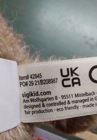 Étiquette d'un jouet en peluche en tissu beige, avec une texture tissée et un ruban blanc portant du texte. Inclut les détails du produit et des notes sur l'éco-responsabilité.