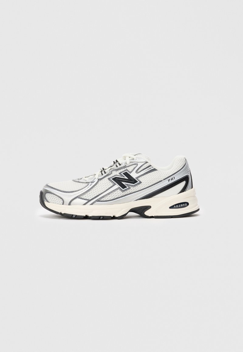 Baskets de sport blanches et grises avec un logo "N" noir, tige en maille, semelle rembourrée et design à lacets, présentées sur un fond gris clair uni.