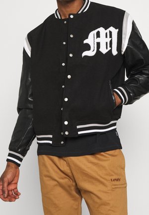 Personne portant une veste varsity noire avec des poignets rayés blancs et un "M" brodé sur la poitrine, associée à un pantalon Levi's beige.