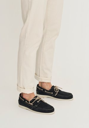 Voeten met zwarte suède mocassin loafers met beige veters, gecombineerd met opgerolde gebroken witte broeken op een lichte achtergrond.