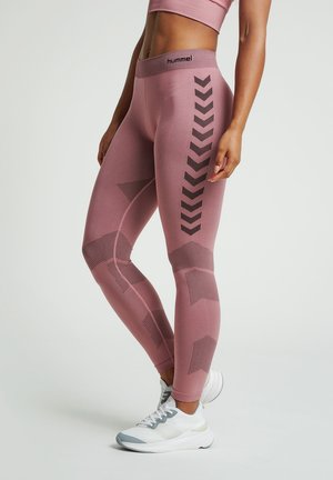 Donna che indossa leggings rosa senza cuciture con motivi geometrici neri e scarpe sportive bianche, in piedi davanti a uno sfondo semplice.