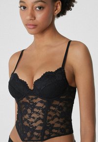 Schwarzer Spitzen-Body mit Unterdraht-Cups, gewellten Kanten und floralem Muster. Verfügt über verstellbare Träger und ein figurnahes Design.