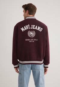 Burgunderfarbene Collegejacke mit weißem Text und Logo auf dem Rücken, mit gestreiftem Rippkragen und -bündchen, weichem Material und sportlichem Design.