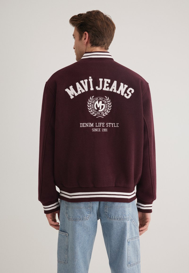 Burgunderfarbene Collegejacke mit weißem Text und Logo auf dem Rücken, mit gestreiftem Rippkragen und -bündchen, weichem Material und sportlichem Design.