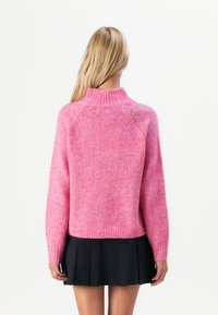 Mulher com cabelo loiro longo a usar um suéter de malha cor de rosa e uma saia preta plissada, de pé, de costas, sobre um fundo branco.