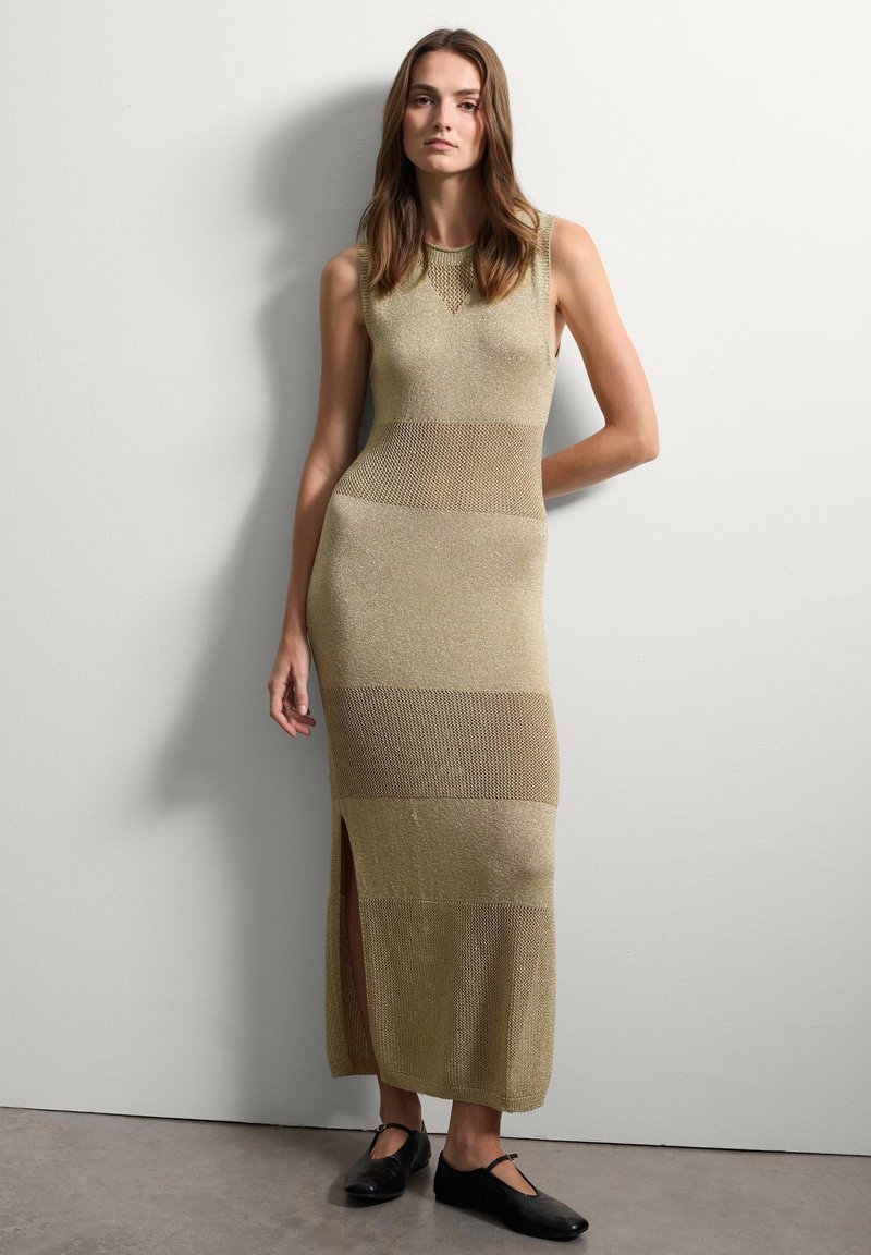 Ärmeloses Strickkleid in gedämpftem Gold mit horizontalem Streifenmuster und seitlichem Schlitz, kombiniert mit schwarzen Ballerinas. Glattes, leichtes Material.