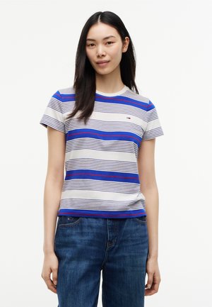 Ung kvinde med langt mørkt hår iført en hvid t-shirt med blå og røde vandrette striber og blå jeans mod en hvid baggrund.