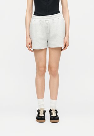 Άτομο φοράει ανοιχτογκρι σορτς με κουμπιά μπροστά και ελαστική μέση, μαύρο τοπ, άσπρες κάλτσες και μαύρα αθλητικά παπούτσια με μπεζ κορδόνια, μπροστά σε λευκό φόντο.