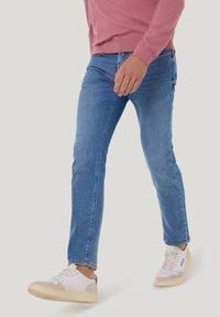 Blaue Denim-Jeans mit Slim Fit und dehnbarem Material, ausgestattet mit einem Five-Pocket-Design, sanften Waschungen und mittlerer Leibhöhe. Kombiniert mit weißen Sneakers.