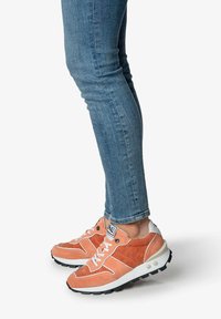 Orange Wildleder-Sneaker mit weißen Schnürsenkeln und Akzenten, die über ein strukturiertes Design und schwarze Gummisohlen verfügen. Tragen Sie sie mit blauen Jeans.