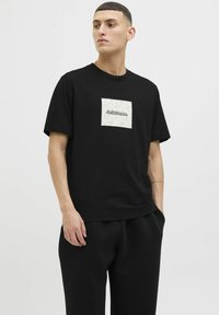 Sort kortærmet T-shirt lavet af bomuld, med en firkantet flødefarvet lap med et blomstermønster og "Jack & Jones" logoet i midten.