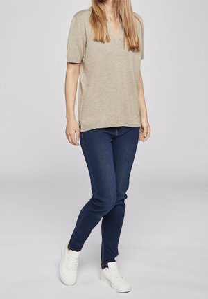 Frau trägt ein beige kurzärmeliges Oberteil, dunkelblaue Skinny-Jeans und weiße Sneakers, steht mit einem Bein leicht angewinkelt.