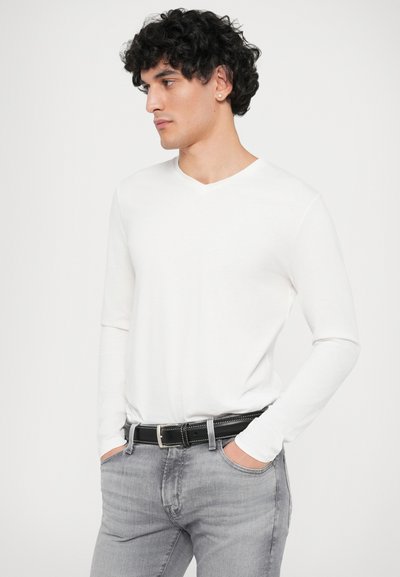 Weißes langärmliges V-Neck-Shirt, kombiniert mit grauen Jeans. Das Modell trägt einen schwarzen Gürtel mit silberner Schnalle. Glattes Material mit minimaler Textur.