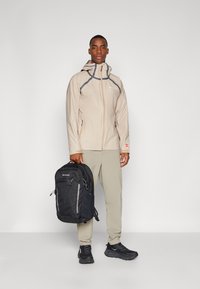 Beige vandtæt jakke med grå detaljer, parret med lysebrune bukser og sorte sneakers, mens der bæres en sort rygsæk.