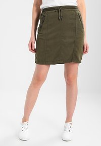 Jupe vert olive avec une taille élastique, cordon de serrage et deux poches zippées. Le design est ajusté avec des détails de couture subtils.