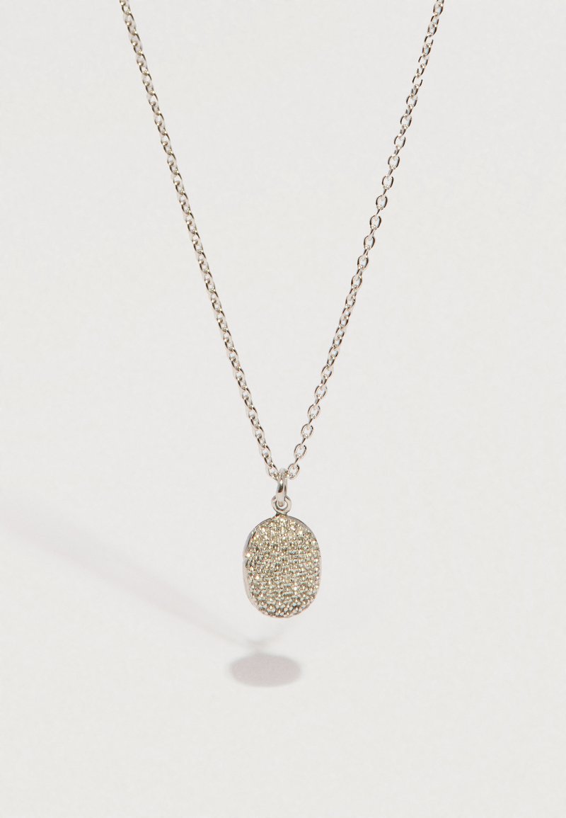 Ασημένιο κολιέ με αλυσίδα και κρεμαστό οβάλ με υφή, ρίχνοντας απαλή σκιά σε ανοιχτό φόντο.