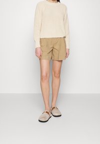 Krémový pletený svetr, béžové šortky s sklady a béžové slip-on boty s hnědými akcenty. Texturovaný materiál a uvolněný střih.