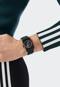 Montre carrée noire avec un bracelet texturé vert. Le cadran de la montre est noir avec une aiguille des heures argentée et un design minimaliste. Manche rayé vert et blanc visible.