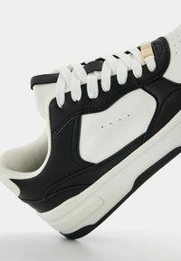 Black and white sneaker featuring a smooth leather upper, textured rubber sole, white laces, and gold accent on the lace eyelet.