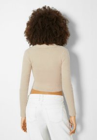 Beige kortärmad tröja med ribbad textur och långa ärmar, med en mjuk krage och fransade kanter, i kombination med vita högmidjade byxor.