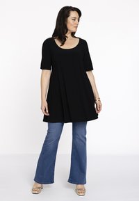 Sort, kortærmet tunika med rund hals og flagrende design, parret med blå flagrende jeans og klare sandalsko med åbne tæer.