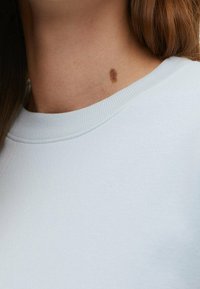 Sudadera azul claro con cuello redondo clásico y textura acanalada, que presenta una pequeña mancha marrón en la zona del cuello.