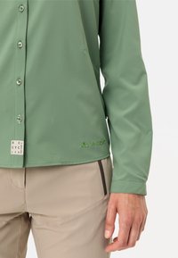 Camicia verde a maniche lunghe realizzata in tessuto leggero con una texture liscia, dotata di chiusura con bottoni e un logo ricamato.
