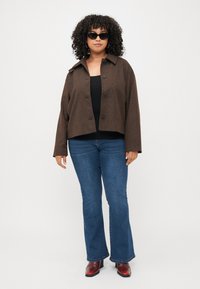 Brown cropped jacket with a collar and buttons, worn over a black top, paired with blue flared jeans and red heeled boots. Black sunglasses worn.