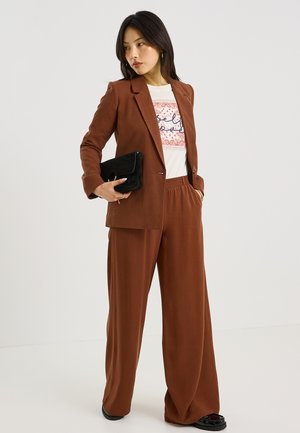 Mujer con traje color óxido con pantalones de pierna ancha, camiseta blanca con estampado, bolso de mano negro y zapatos negros, de pie sobre un fondo blanco.