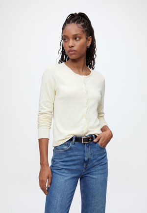 Donna con i capelli intrecciati che indossa un cardigan crema con bottoni e jeans blu con cintura nera, in piedi con una mano in tasca su sfondo bianco.