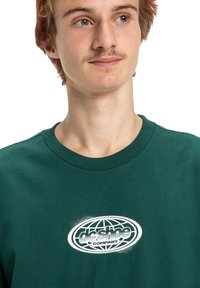 Młody mężczyzna z jasnobrązowymi włosami i wąsem, ubrany w ciemnozieloną bluzę z białym logo "dc shoe company" z wzorem globu.