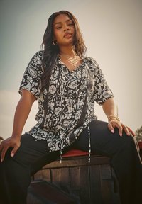 Svart och vit mönstrad kortärmad blus med v-hals och snörningsdetaljer, i kombination med svarta byxor. Slät tygtextur.