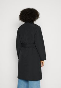 Abrigo largo negro con cinturón, que presenta una textura suave y un diseño minimalista. La parte posterior tiene un dobladillo ligeramente acampanado y mangas amplias.