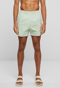 Lys grønne badebukser med elastisk talje og sidelommer. Brugt med beige slidersandaler. Glat tekstur, afslutning ovenfor knæet.