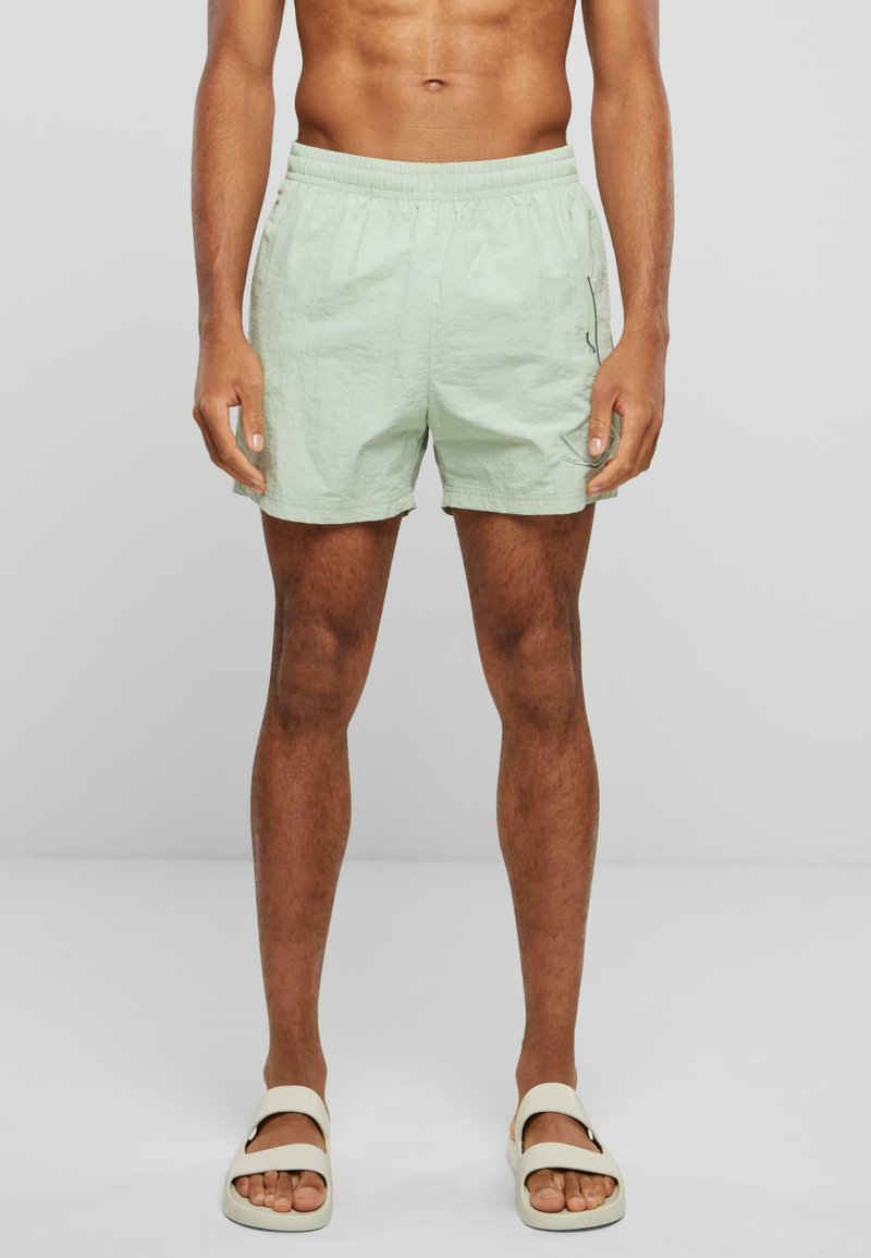 Lys grønne badebukser med elastisk talje og sidelommer. Brugt med beige slidersandaler. Glat tekstur, afslutning ovenfor knæet.