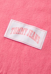 Ružová tkanina s jemnou textúrou má bielu nálepku s nápisom "TOMMY JEANS" v ružových blokových písmenách, zakrivenú hore.