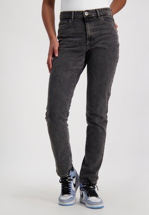 Personne portant un jean noir délavé taille haute et des baskets montantes blanches et bleues, debout devant un fond clair uni.