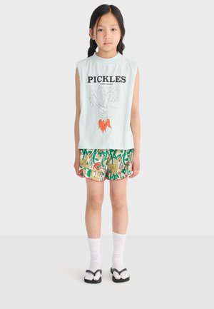 Young girl stands wearing a sleeveless shirt with "PICKLES" text and vegetable illustration, floral shorts, white socks, and black sandals.