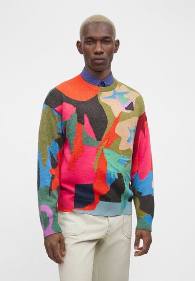 Άνδρας με κοντά ξανθά μαλλιά που φοράει φωτεινό πουλόβερ με πολύχρωμο αφηρημένο σχέδιο και μπεζ παντελόνια, στέκεται σε απλό φόντο.